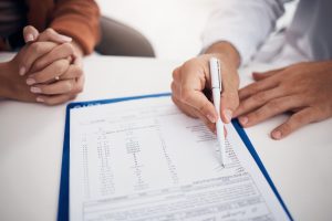Doctor, hands and writing on documents with patient for life insurance, policy or legal agreement at hospital. Closeup of medical employee showing paperwork, agreement or checklist on desk at clinic