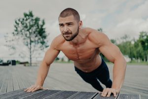 Shirtless bearded man does push-ups in park, trains upper body, maintains regular workout routine. Motivated athlete in action.